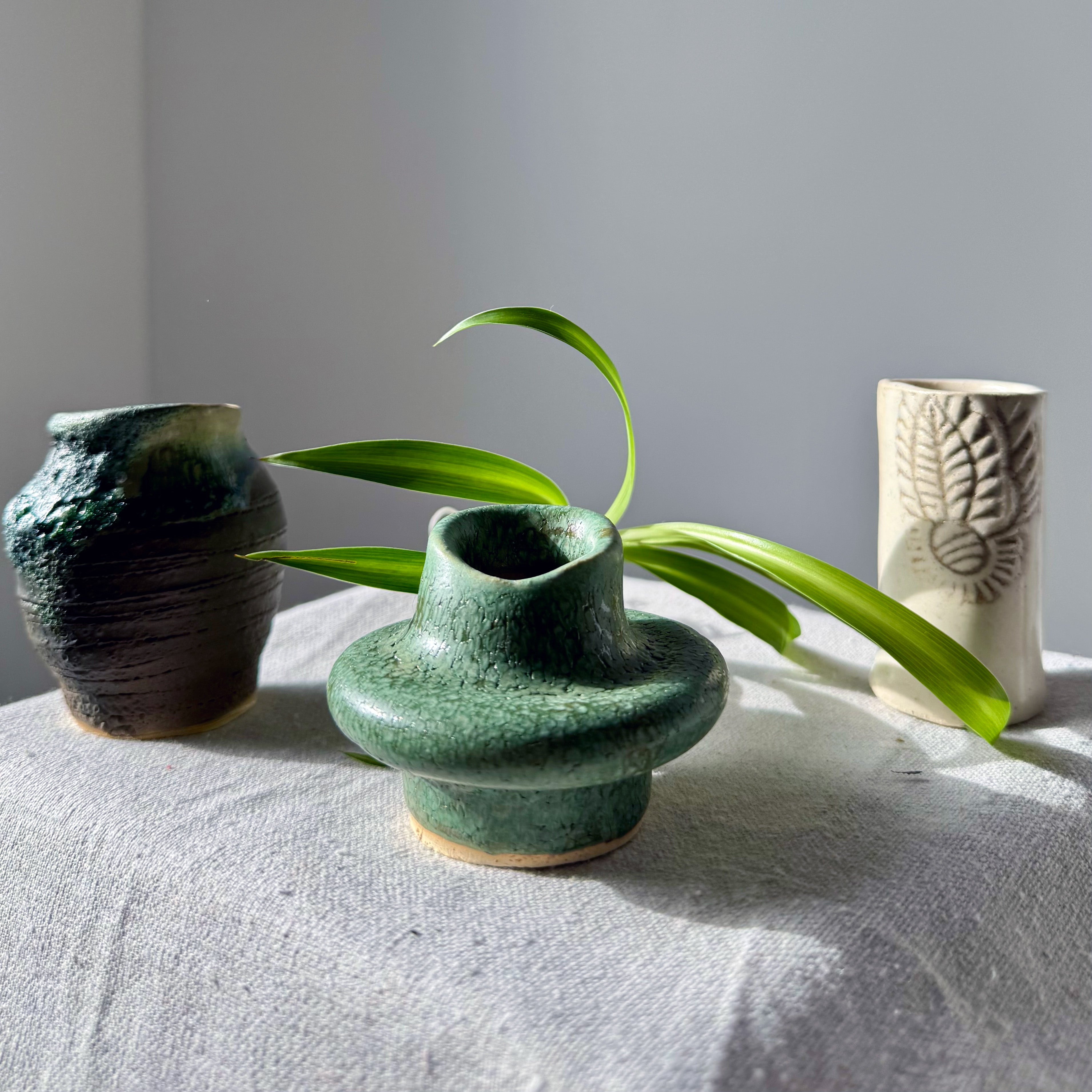 Three ceramic vases on a textured surface with a plant leaf