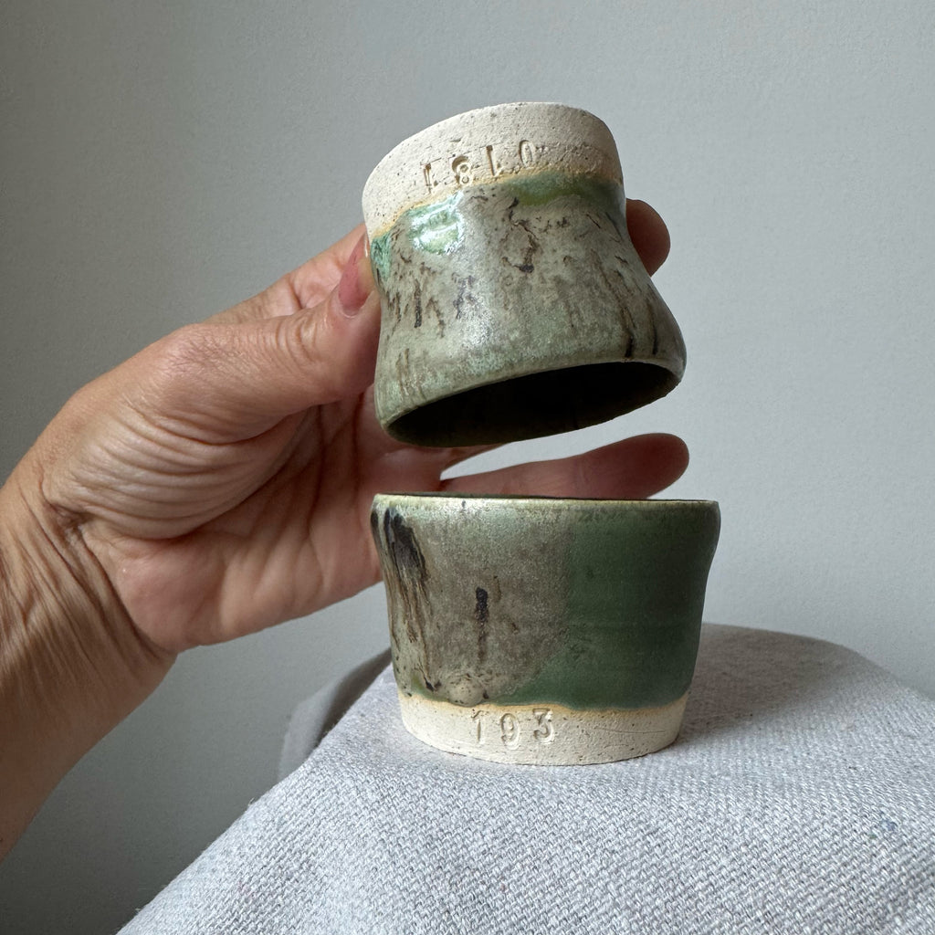 Hand holding two ceramic cups with green glaze against a neutral background