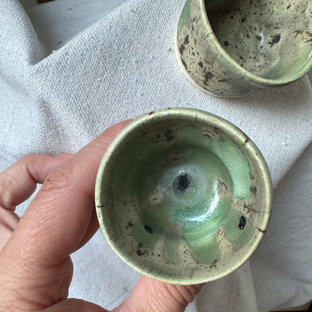 Hand holding a green ceramic cup with another cup in the background on a textured surface.