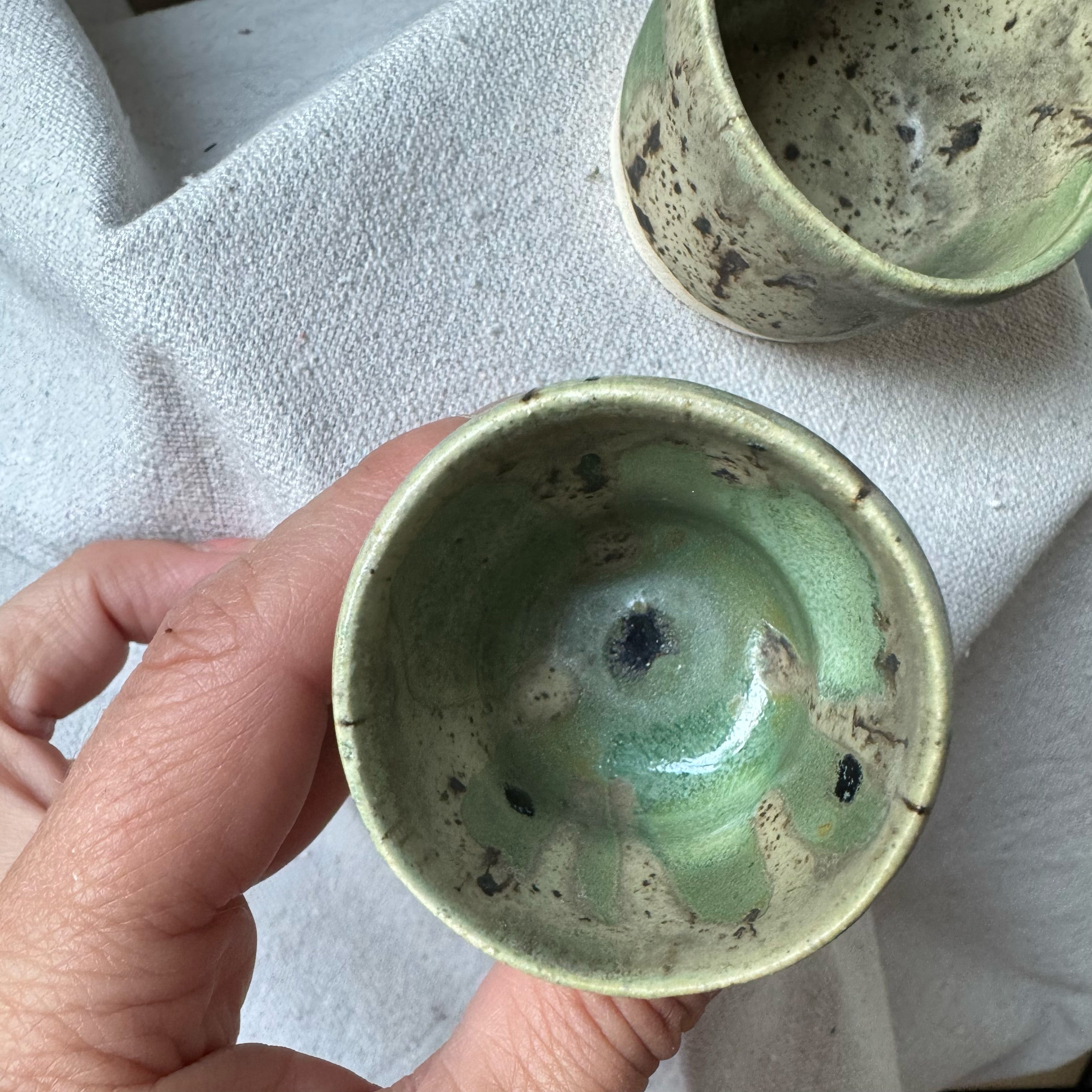 Hand holding a green ceramic cup with another cup in the background on a textured surface.