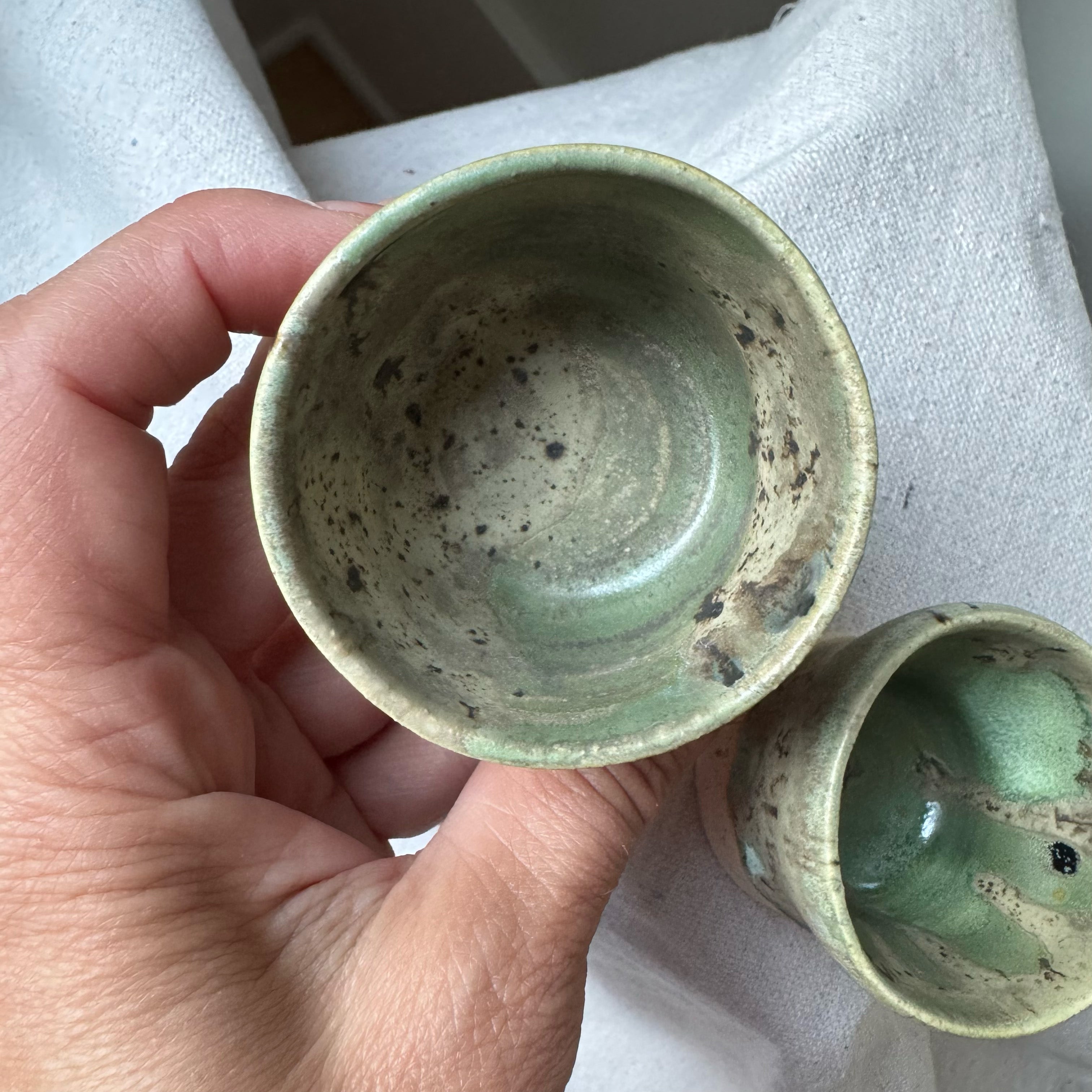 Hand holding a green ceramic cup with another cup in the background on a light surface.
