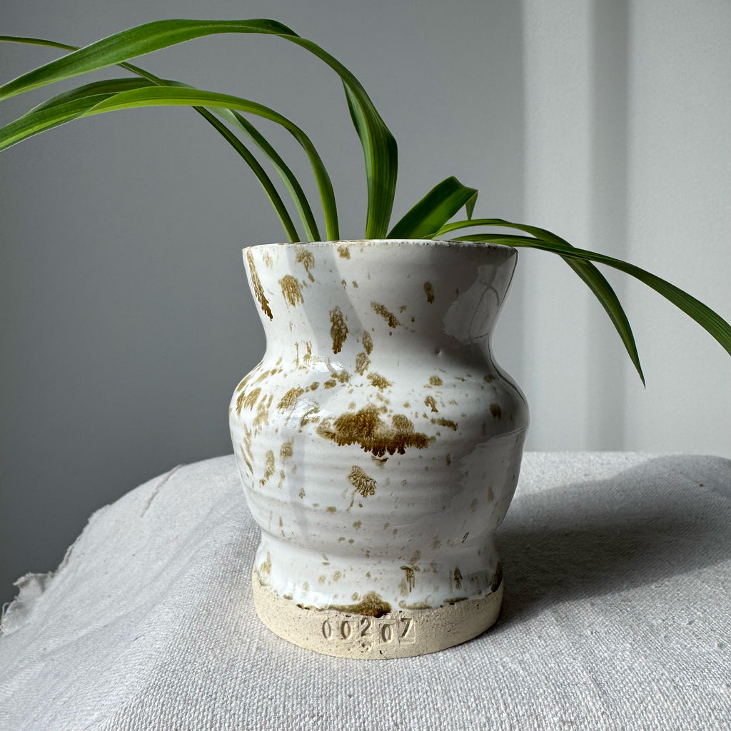 White ceramic vase with gold speckles containing a plant on a light surface with a neutral background