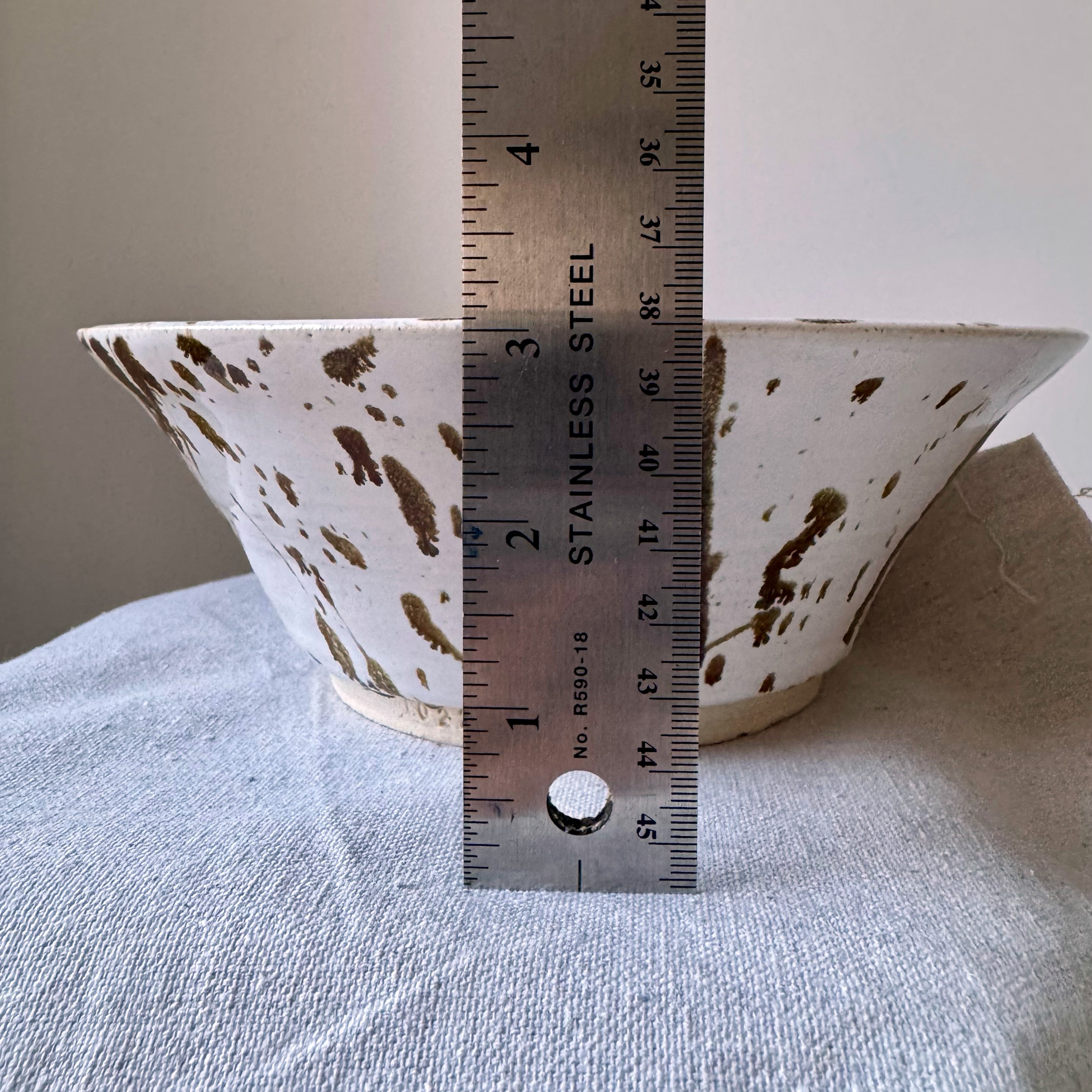 Ceramic bowl with brown speckles and a ruler for scale on a textured surface.