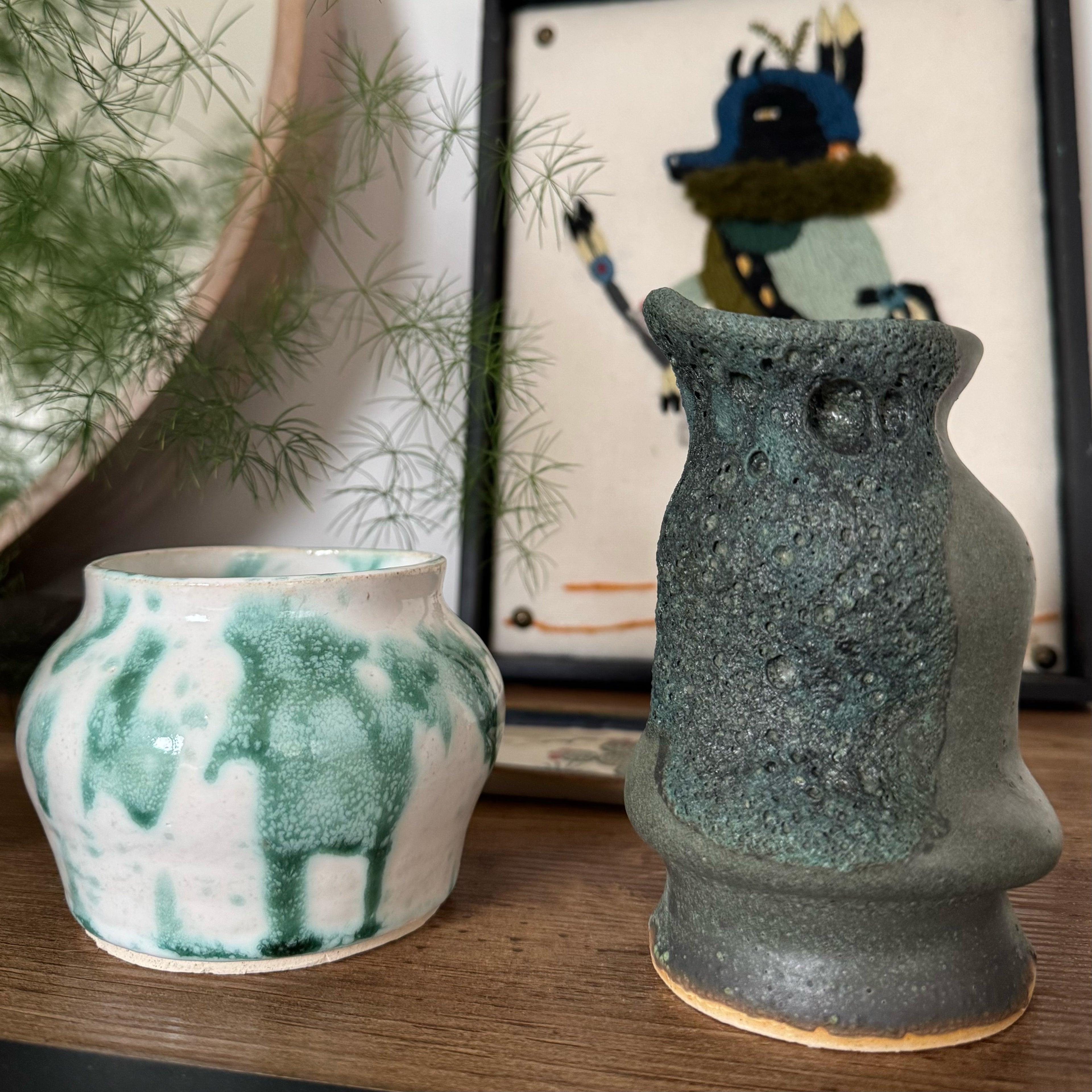 Two ceramic vases on a wooden surface with a plant and framed picture in the background.