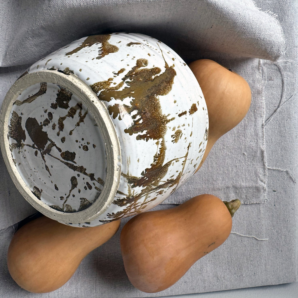 White ceramic pot with brown abstract patterns on a gray surface with two gourds.