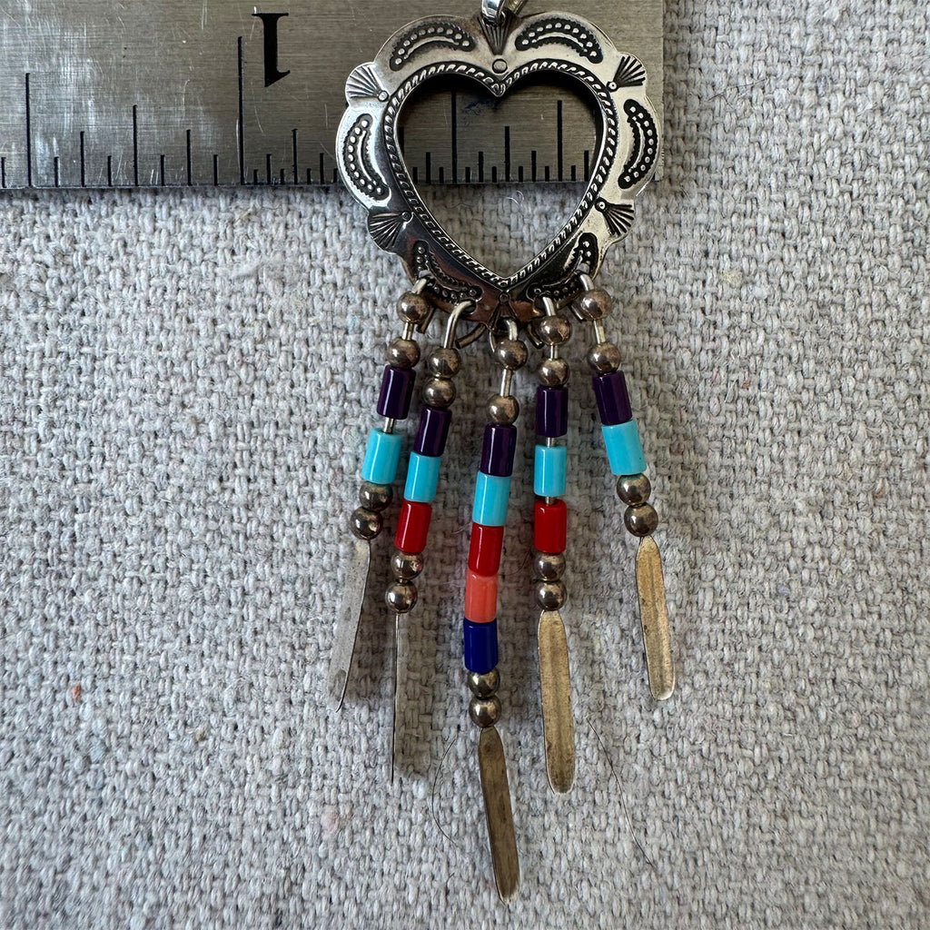 Decorative heart-shaped pendant with beads and feathers on a textured surface with a ruler for scale.