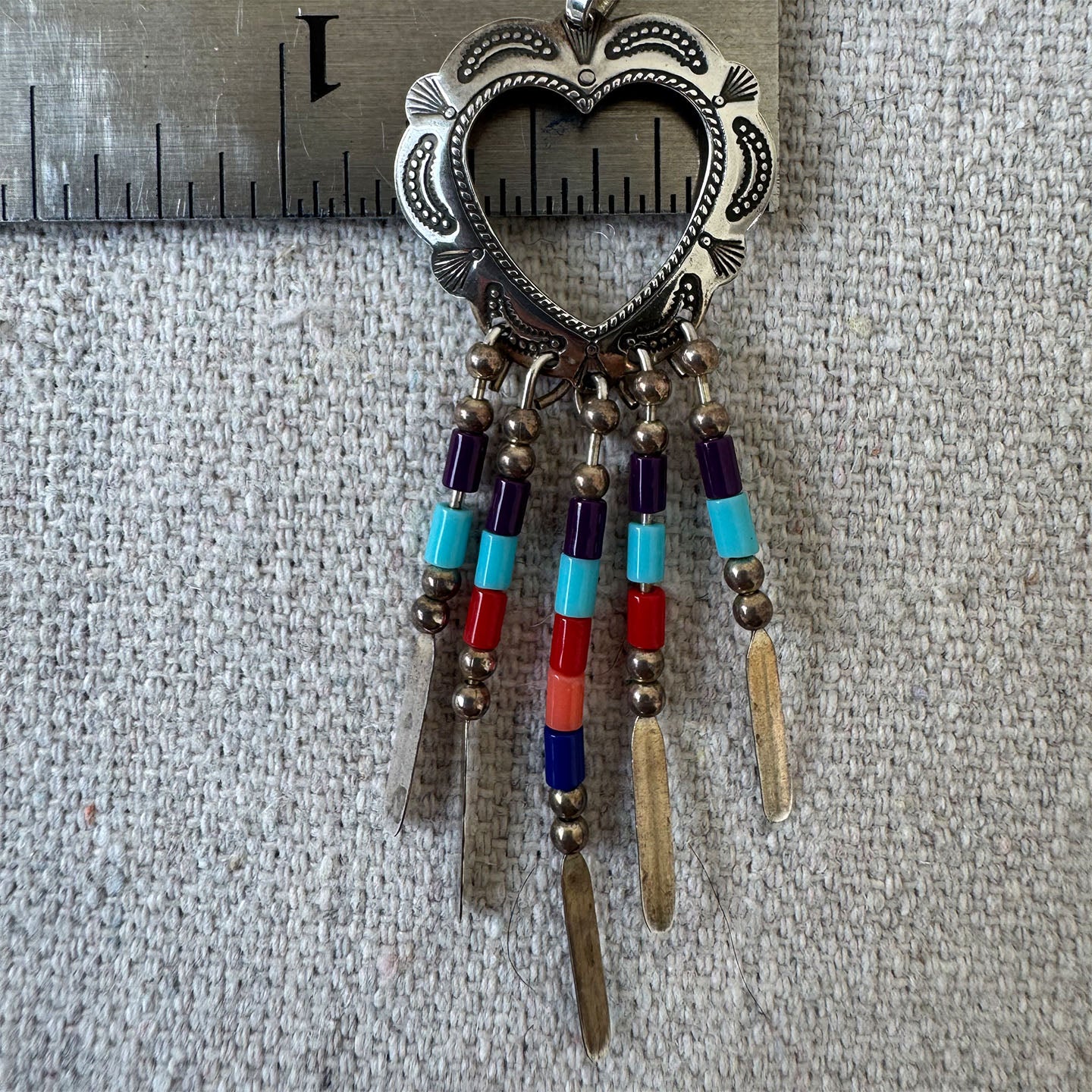 Decorative heart-shaped pendant with beads and feathers on a textured surface with a ruler for scale.