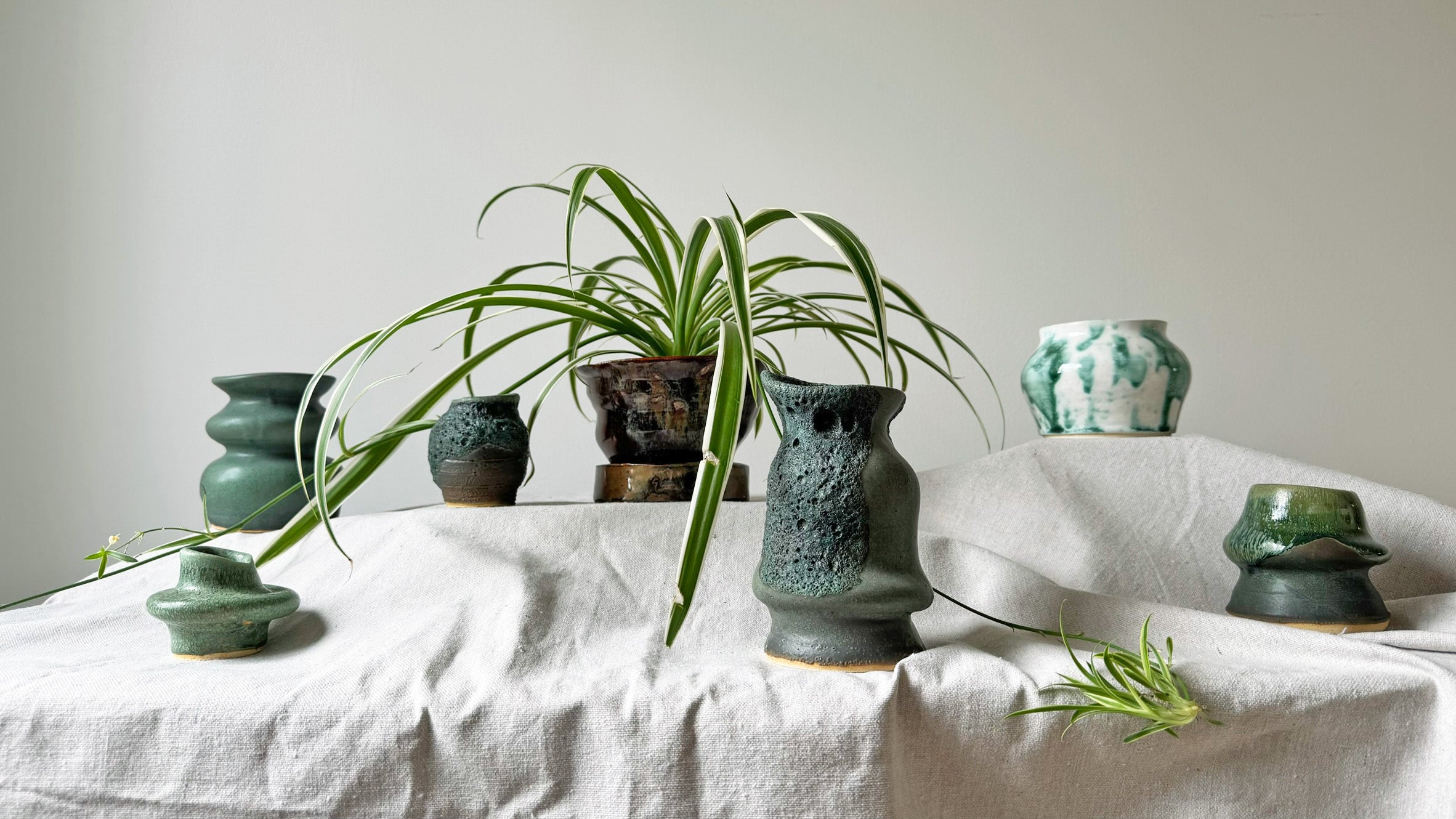 Green ceramic vases and plants on a white tablecloth against a plain wall